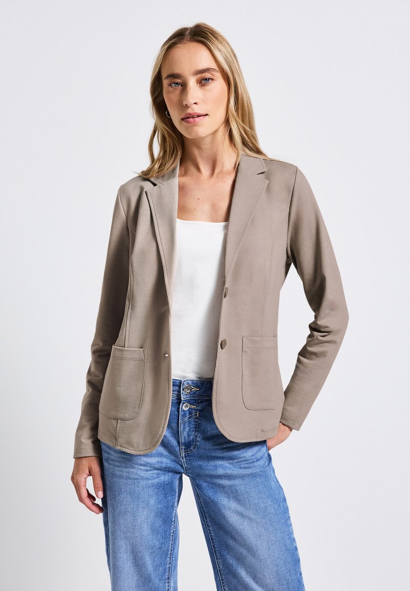 Beige lättviktsblazer med två framfickor, notchade slag och knäppning, ihopparad med en vit topp och blå jeans.