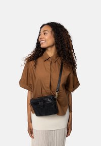 Giovane donna che sorride e guarda di lato, indossando una maglietta marrone a maniche corte, una gonna color crema e una borsa a tracolla in pelle intrecciata nera.