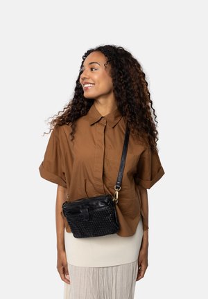 Jonge vrouw die glimlacht en opzij kijkt, gekleed in een bruine shirt met korte mouwen, een crèmekleurige rok en een zwarte geweven leren schoudertas.