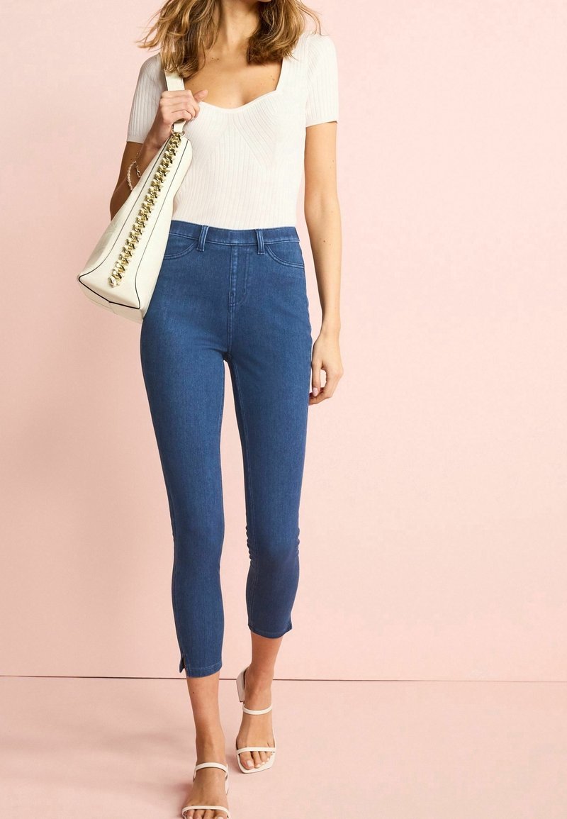 Femme portant un haut blanc à manches courtes, un jean bleu taille haute raccourci, des sandales blanches, et un sac à main blanc avec un détail de chaîne dorée.