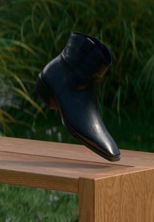 Bottine noire en cuir avec un petit talon flottant au-dessus d'un banc en bois en plein air avec un feuillage vert en arrière-plan.
