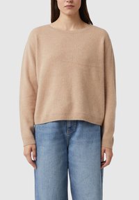 Beżowy dziany sweter o luźnym kroju, z okrągłym dekoltem i długimi rękawami. Miękka tekstura, prążkowane mankiety i dół. Połączony z jasnoniebieskimi dżinsami.