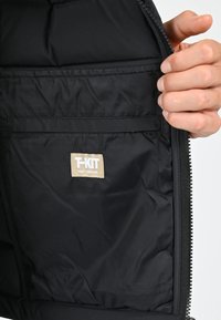 Intérieur de veste noire avec une texture lisse, fermeture éclair et une étiquette beige avec le texte "T-KIT". Comprend une grande poche.
