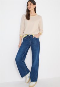 Pull beige clair avec des poignets jaune néon, associé à un jean large bleu foncé et des baskets beige avec des accents bleu clair.