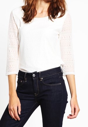 Donna che indossa un top bianco in pizzo a maniche lunghe e jeans blu scuro, in piedi con una mano vicino all'anca e l'altra rilassata.