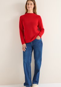 Czerwony sweter z prążkowanym wzorem i długimi rękawami, w zestawieniu z niebieskimi dżinsami. Model stoi na neutralnym tle, ręce w kieszeniach.
