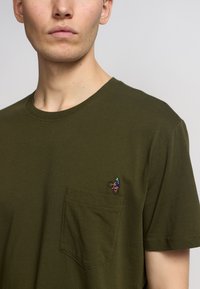 Camiseta de algodón verde oliva con un bolsillo en el pecho y un pequeño diseño bordado de una persona en bicicleta. Manga corta, escote en cuello redondo.