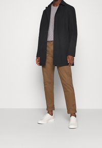 Svart jacka med spetsig krage, enkel knappknäppning, bärs över en randig skjorta. Matchas med bruna byxor och vita sneakers.