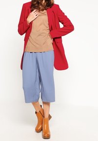Röd kavaj över en beige tröja, matchad med ljusblå culotter och bruna ankelstövlar. Kavajen har en strukturerad design och fickor.