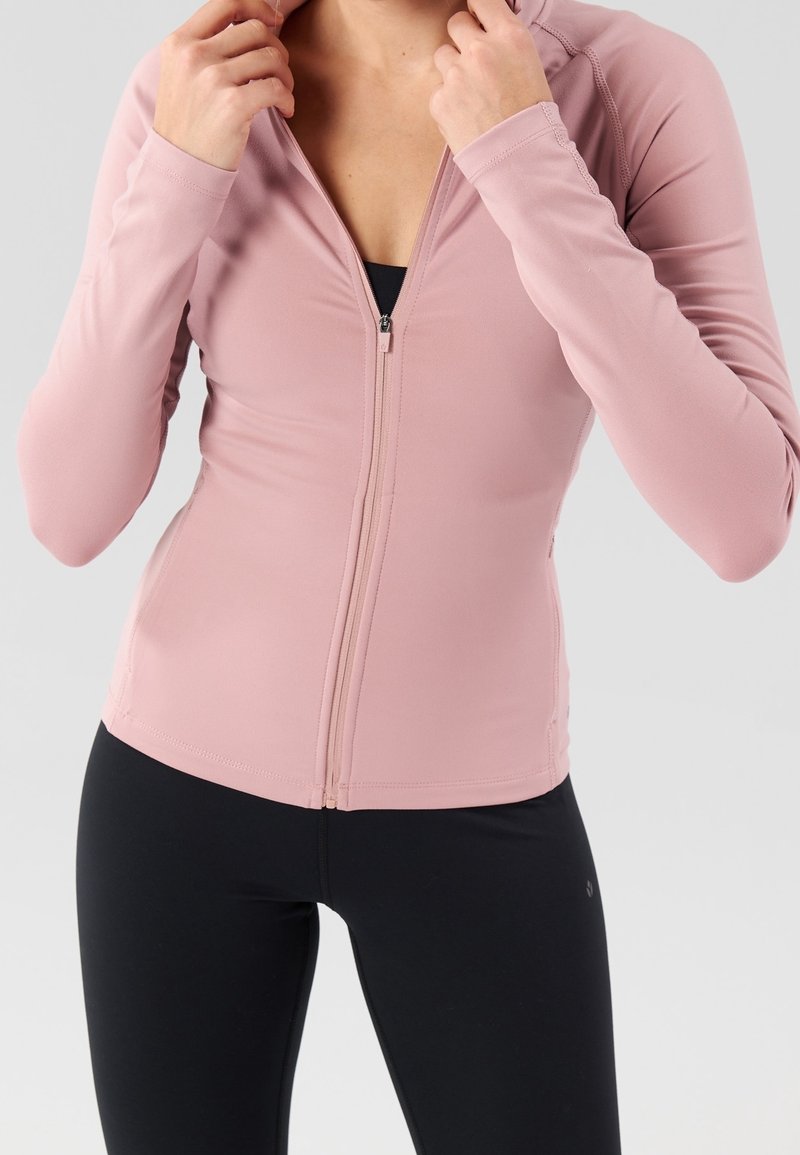 Femme portant une veste zippée rose clair par-dessus un haut noir et des leggings noirs, ajustant le col de sa veste avec ses deux mains.