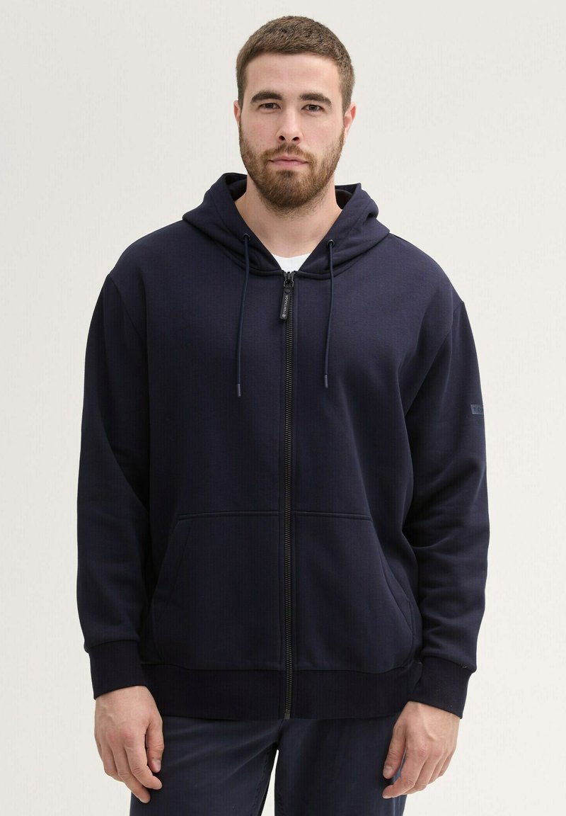 Marineblauwe zip-up hoodie van gladde stof, met een capuchon met trekkoord, voorzakken en ribbelboorden. Eenvoudig, praktisch ontwerp.