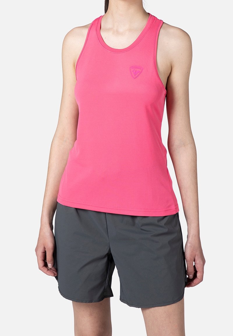 Žena v růžovém sportovním tílku bez rukávů a volných tmavě šedých šortkách, stojící na pozadí s jednoduchým světlým pozadím.