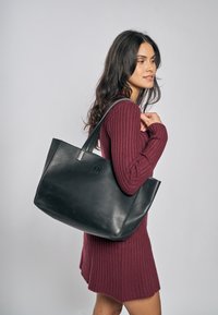 Borsa tote in pelle nera dal design strutturato con manici lunghi, mostrata accanto a una modella che indossa un vestito a coste color bordeaux.