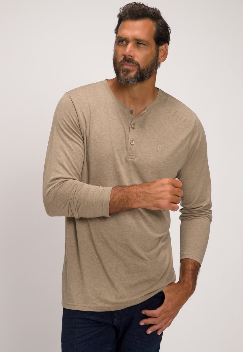 Beige Langarm-Henley-Shirt mit drei Knöpfen und weichem Material. Mit dezentem Logo auf der Brust und figurbetonten Ärmeln. Getragen mit dunklen Jeans.