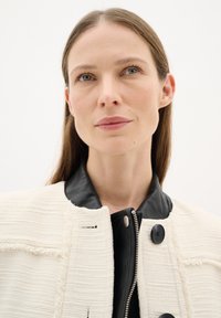 Giacca bianca con texture e colletto in pelle nera, con chiusura a zip e bottoni neri rotondi. Il tessuto liscio contrasta con il motivo lavorato a maglia.