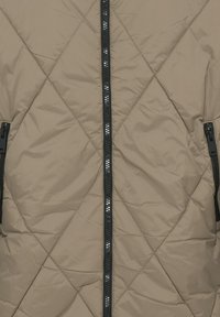 Beige gesteppte Jacke mit Rautenmuster und schwarzem Reißverschluss, der senkrecht in der Mitte verläuft, mit zwei seitlichen Reißverschlusstaschen.