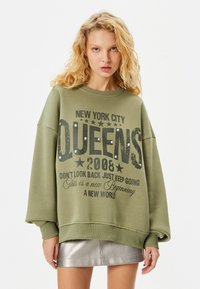 Mujer rubia vestida con una sudadera oversized verde oliva que dice "New York City Queens 2008", con tachuelas y una falda mini plateada, sobre un fondo blanco.