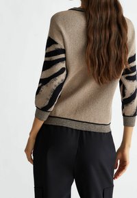 Pull en maille beige avec fil métallique, présentant des rayures abstraites noires sur les manches et un ourlet et des poignets côtelés.