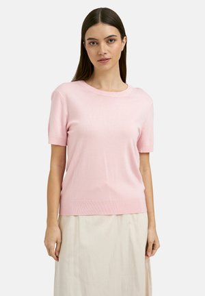 Woman with straight dark hair wearing a light pink short-sleeve knit top and a light beige skirt standing against a plain background.