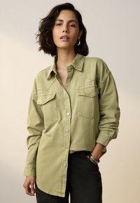 Grünes Denimhemd mit Knopfleiste, zwei Brusttaschen und langen Ärmeln. Der Stoff hat eine strukturierte Oberfläche.