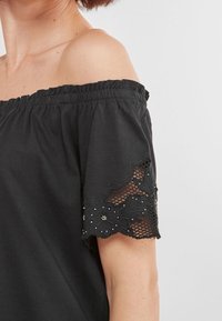Haut noir épaules dénudées avec manches en dentelle texturée ornées de motifs floraux et de petites perles décoratives. Tissu doux, coupe décontractée.