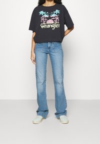 Svart oversized t-shirt med korta ärmar och flerfärgad "Wrangler" grafik, tillsammans med ljusblå flare jeans och mintgröna skor.