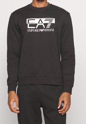 Person trägt schwarzen Sweatshirt und Hose mit weißem Emporio Armani EA7-Logo auf Brust und Hose.
