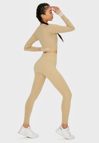 Beige langärmliges Cropped-Top und passende hochtaillierte Leggings, glatte Textur, figurbetontes Design, akzentuiert mit weißen Streifen und Logo.