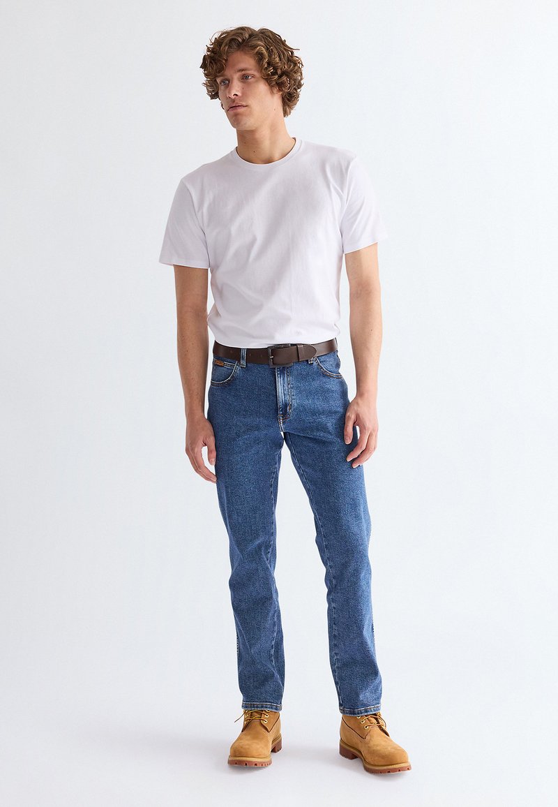 Ung mand, stående, iført en ensfarvet hvid t-shirt, blå jeans med et brunt bælte og tan arbejdsstøvler, mod en hvid baggrund.