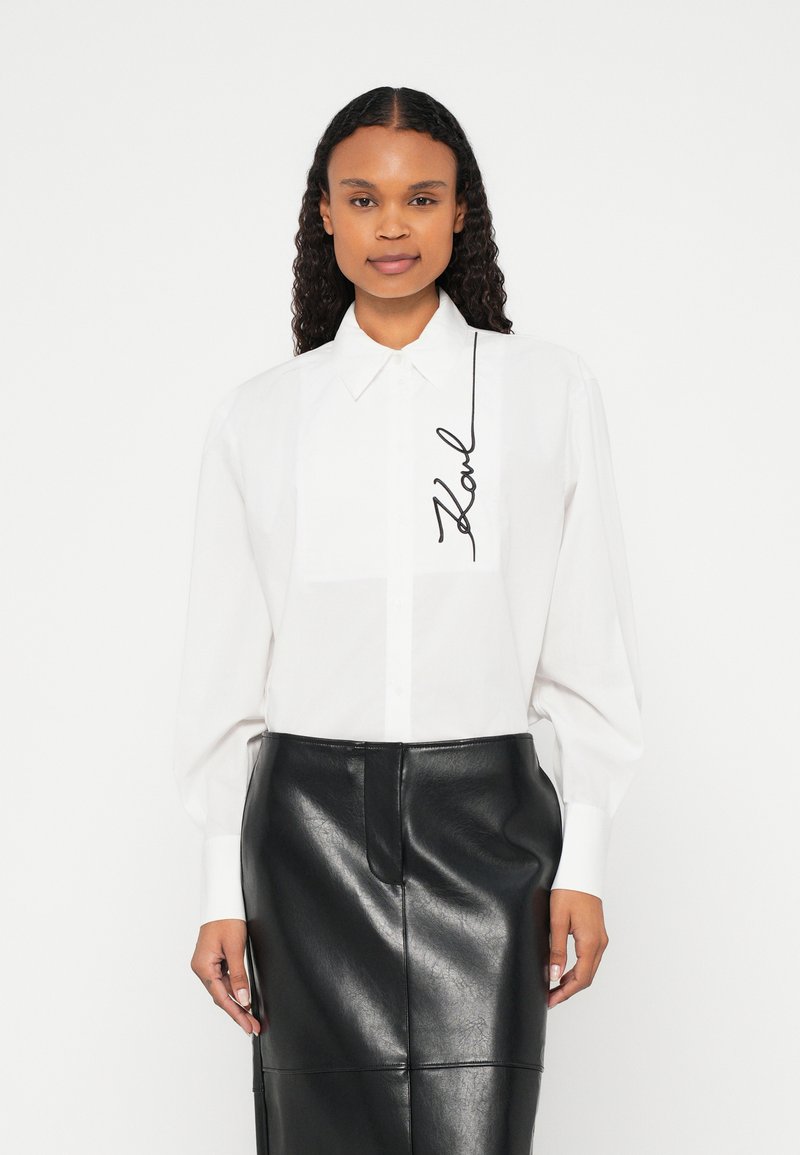 Ženska, oblečena v belo srajco z dolgimi rokavi z vezenim napisom "Karl" in črno usnjeno krilo z visokim pasom, stoji pred belim ozadjem.