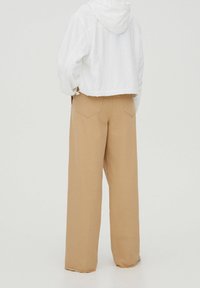 Bílá krátká bunda s kapucí a stahovací šňůrkou, kombinovaná s béžovými širokými kalhotami s kapsami na zadní straně a třepenými lemy.