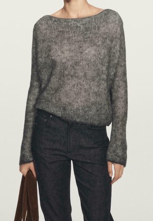 Personne portant un pull en maille gris ample à manches longues et un jean taille haute foncé, tenant un objet en tissu marron dans une main.