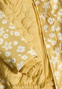 Giacca imbottita gialla con motivi floreali bianchi, chiusura con zip e polsini elasticizzati. Texture morbida e design leggero.