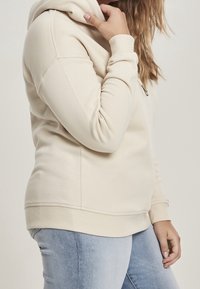 Femme portant un sweat à capuche beige clair avec détail zippé, tenant la capuche près du cou, vêtue d'un jean bleu clair sur un fond uni.