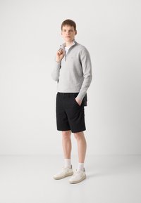 Světle šedý pletený pulovr s polo zipem, černé šortky a krémové tenisky s bílými ponožkami; moderní volnočasový outfit s uvolněným střihem.