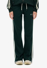Grüne Sweatpants aus weichem Stoff, mit weißen Seitenstreifen und einem elastischen Bund mit Kordelzug. "ATHLETE" auf der Vorderseite aufgedruckt.