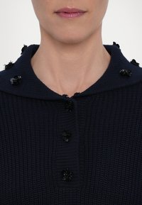 Gros plan sur le bas du visage et le cou d’une personne portant un pull en maille bleu marine avec des ornements floraux noirs le long du col et de la ligne des boutons.