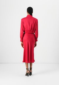Robe rouge vif avec des manches longues et un design court, présentant une taille élastique et un tissu lisse et fluide. Talons noirs à lanières.