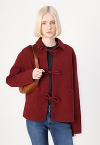 Burgundska jakna s ovratnikom i mašnom, s džepom, u kombinaciji s smeškom teksturiranom torbicom i plavim farmerkama.