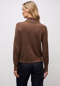 Brauner Strickpullover mit hohem Kragen, gerippten Bündchen und Saum, präsentiert an einem Modell in dunkler Jeans. Glatte Textur, taillierte Passform.