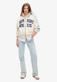 Chaqueta cremosa con cremallera y texto bordado en azul "SUPERDRY ATHLETIC" con acentos a rayas azules. Combinada con jeans azul claro y zapatillas blancas.