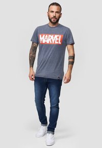 Šedé tričko s velkým červeným a bílým logem "MARVEL". Nosené s modrými džíny a bílými teniskami. Ležérní střih a krátké rukávy.