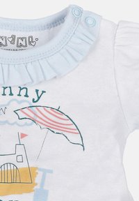 Valkoinen lyhythihainen t-paita, jossa on vaaleansiniset röyhelökaulus. Siinä on värikäs printti, jossa on sateenvarjo, linna ja aaltoja pastelliväreissä.