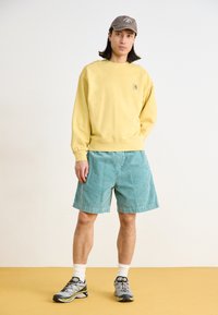 Sweatshirt en polaire jaune, shorts en velours côtelé teal, casquette grise et chaussures de sport grises avec des accents vifs. Porté avec des chaussettes blanches à mi-mollet.
