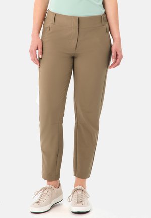 Femme portant un pantalon beige ajusté avec des poches latérales zippées et des baskets beiges à lacets, debout devant un fond uni.
