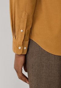 Camisa de corduroy mostaza con puños abotonados, presenta una manga puntiaguda y una sutil textura acanalada, combinada con tejido de cuadros marrón.