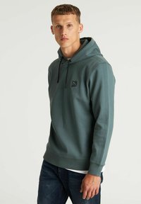 Felpa con cappuccio in tessuto verde con una trama liscia, caratterizzata da un patch logo frontale, lacci neri e dettagli con strisce laterali.