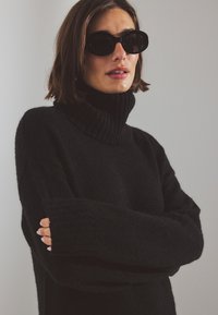 Musta korkea kauluksinen neule, jossa on pehmeä, teksturoitu pinta ja leveät ribbaus yksityiskohdat; yhdistettyinä suuriin pyöreisiin mustiin aurinkolaseihin.