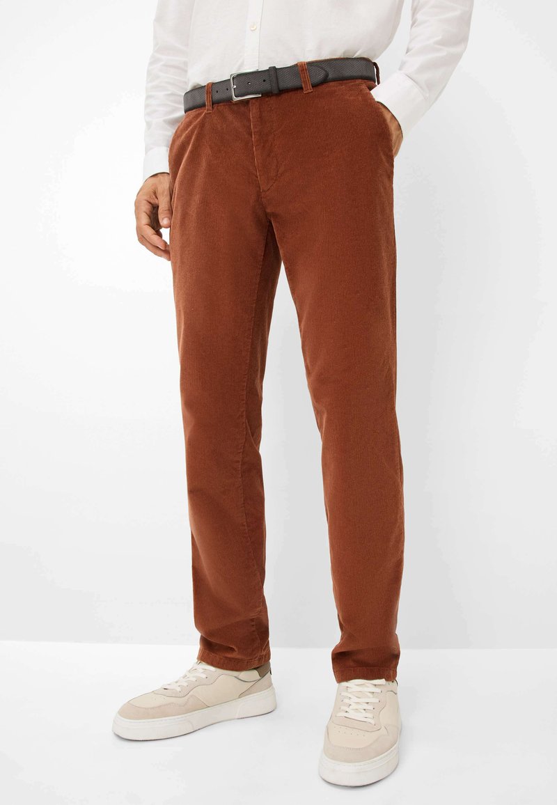 Pantalon en velours côtelé marron avec une coupe droite, équipé d'une ceinture et de poches latérales, associé à des baskets claires sur un fond neutre.