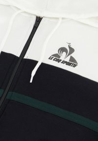 Felpa con zip bianca e nera caratterizzata da dettagli a righe verdi, materiale in cotone, un logo sulla parte superiore sinistra e cappuccio con cordoncino.